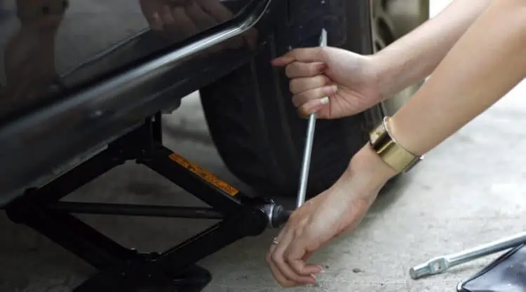 A ladies hands using a black scissor jack, to raise a black car