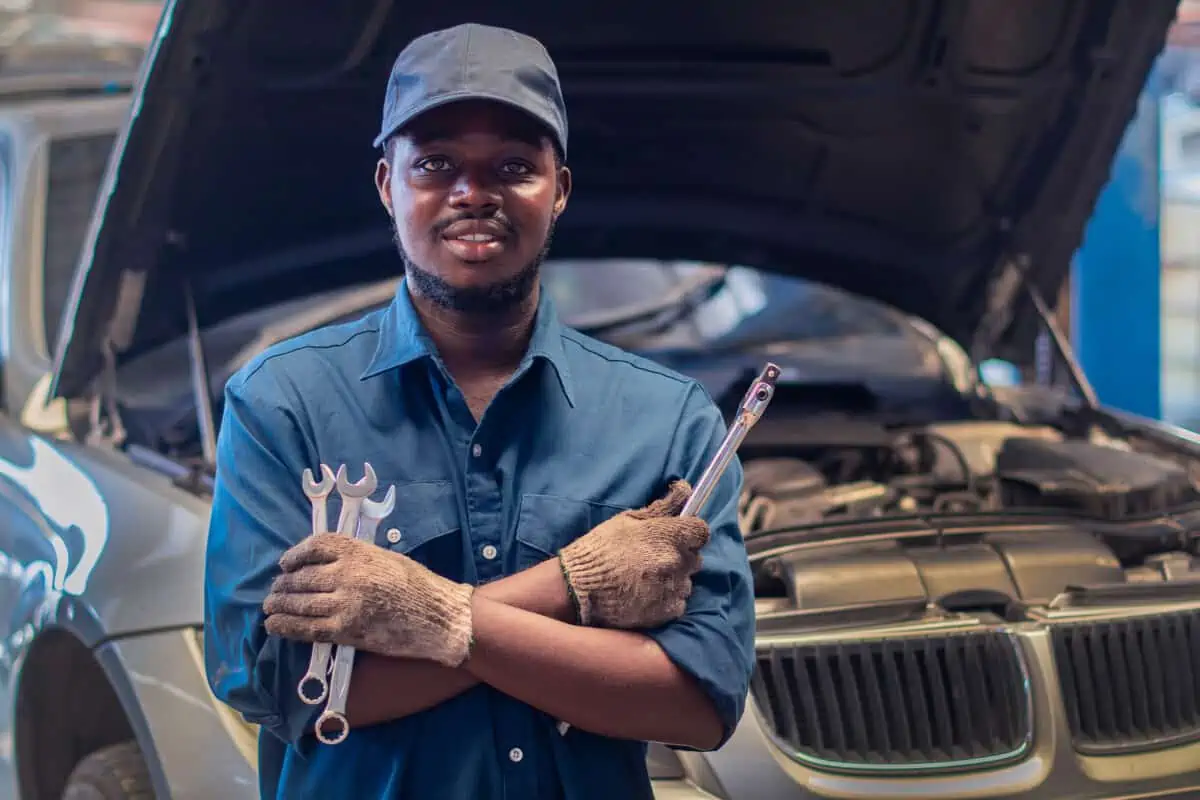 A smiling car mechanic, holding tools in front of a car with the hood open