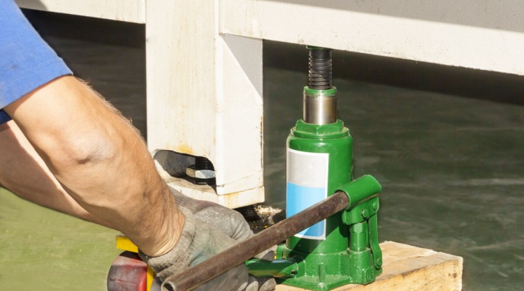 A man using a hydraulic bottle jack to lift what looks like a shipping container