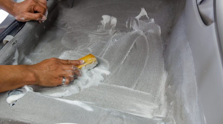 A man scrubbing a cars grey carpet with a yellow scrubbing brush