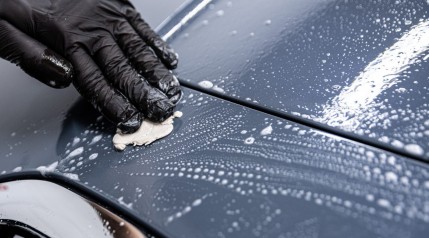 A black gloved hand cleaning a soapy car with a clay bar