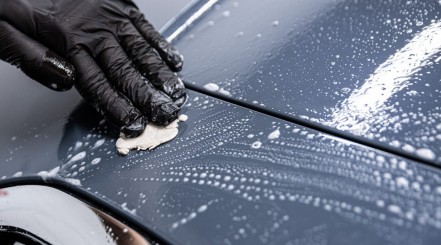 A black gloved hand cleaning a soapy car with a clay bar