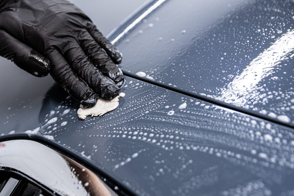 A black gloved hand cleaning a soapy car with a clay bar