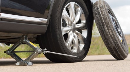 Scissor jack under a black car with a spare wheel beside it